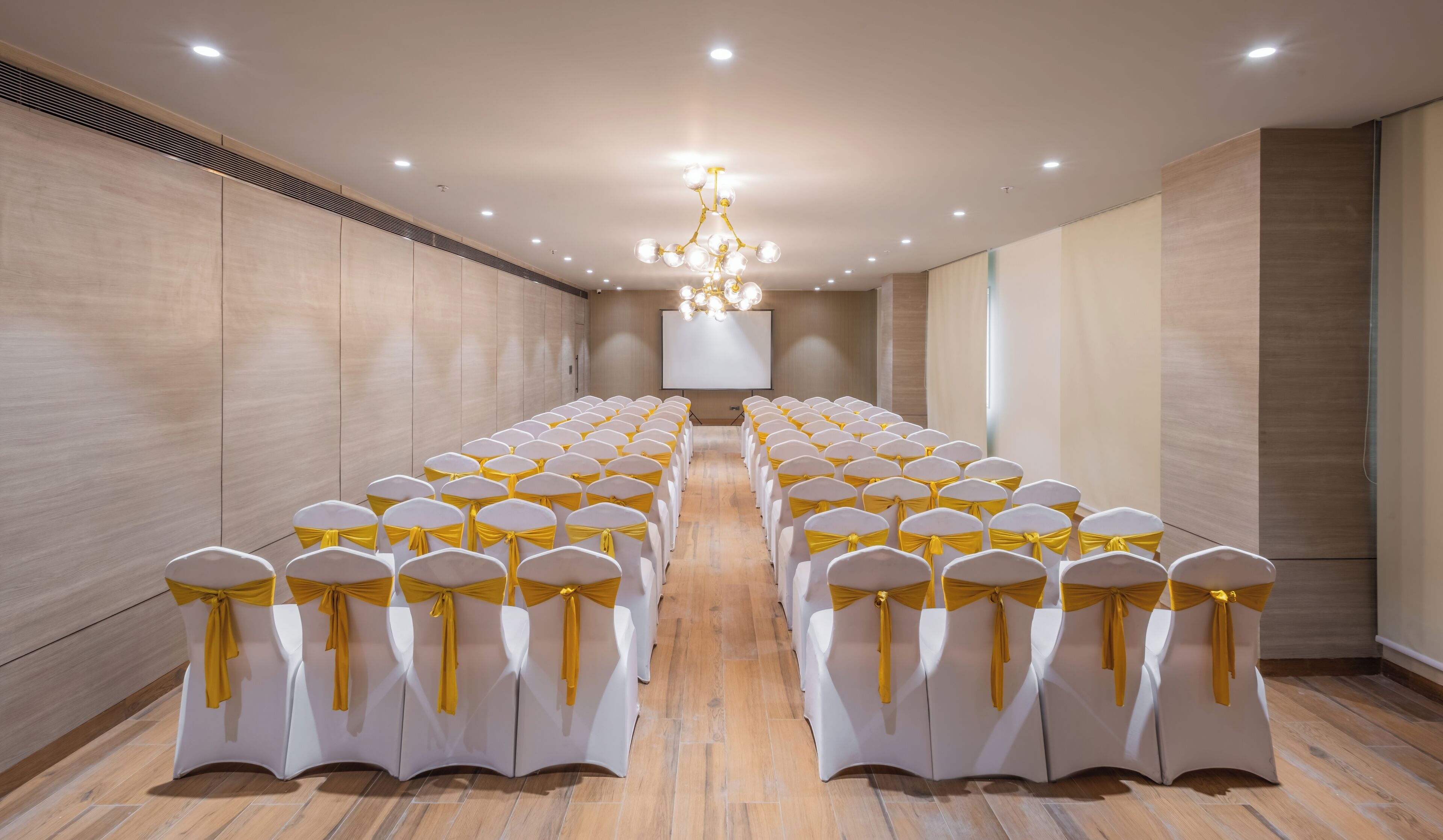 A conference room with rows of white chairs with yellow sashes, a large chandelier, and a projector screen.