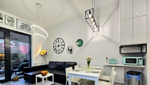 A modern living room with a black leather sofa, a dining table set for four, and a kitchen area with a microwave. - Apartament Zielony Przylądek - 365PAM (Ustronie Morskie)