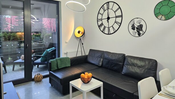 A modern living room with a black leather sofa, a small white table with a bowl of fruit, and decorative wall clocks. - Apartament Zielony Przylądek - 365PAM (Ustronie Morskie)
