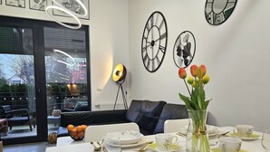 A modern dining area with a table set for two, a sofa, and a large window with blinds. - Apartament Zielony Przylądek - 365PAM (Ustronie Morskie)