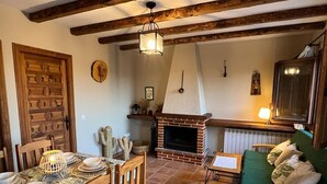 Un salon rustique avec des poutres apparentes, une cheminée en briques, une table à manger dressée avec de l’argenterie et une bougie, et un canapé vert recouvert de tissu.