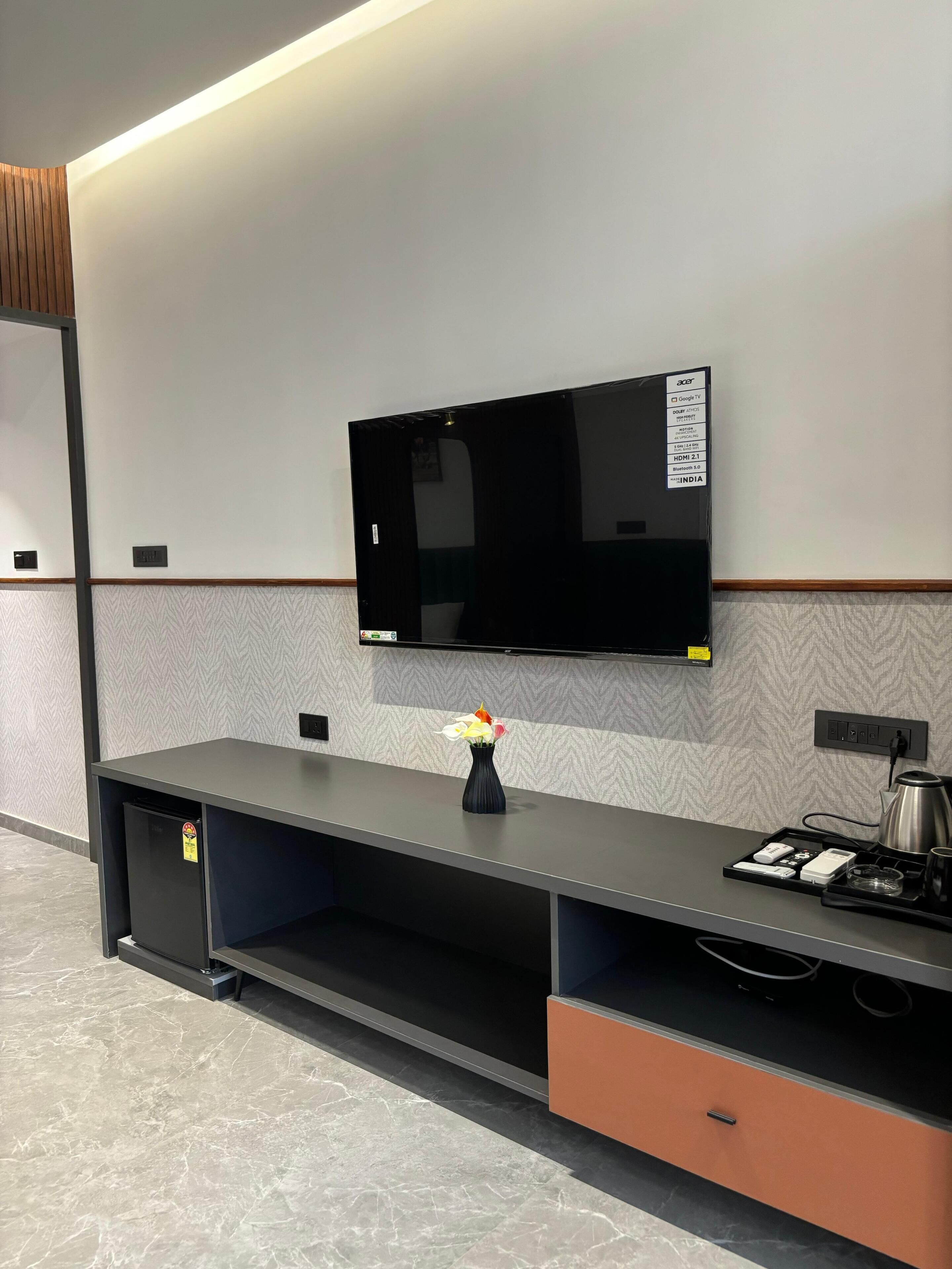 Una habitación de hotel moderna con un televisor de pantalla plana montado en la pared, un pequeño florero sobre la mesita consola, y una cafetera a un lado.