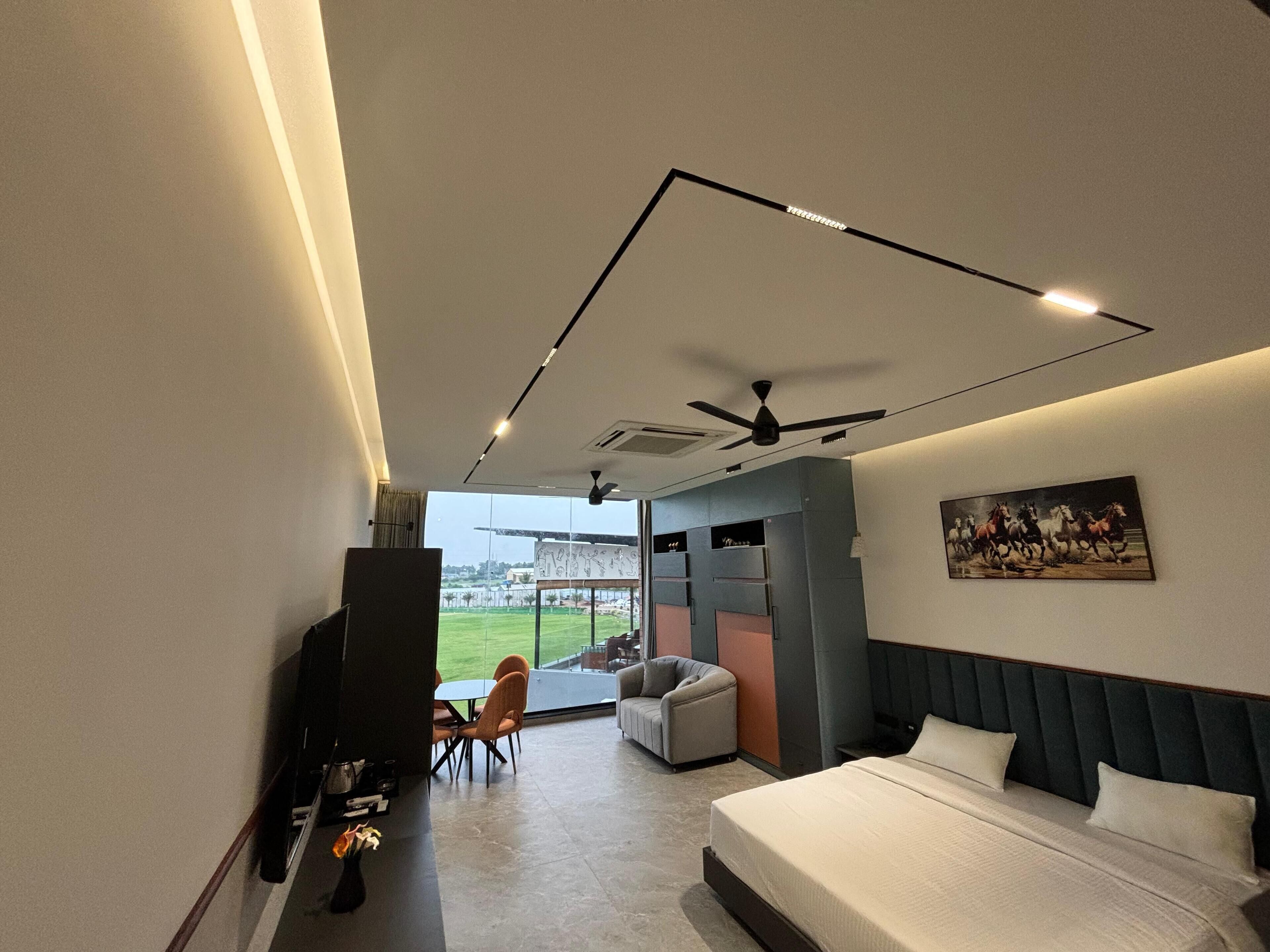 Habitación de hotel moderna con una cama grande, un ventilador de techo, una zona de comedor con sillas y vistas a un paisaje verde a través de amplias ventanas.