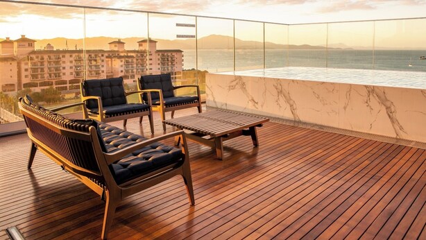 Una terraza de madera con dos sillones de descanso y una mesa, con vista a un cuerpo de agua y edificios a lo lejos.