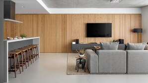 Una sala de estar moderna con un sofá gris, paneles de madera, un televisor de pantalla plana y una zona de cocina con taburetes.