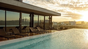 Una piscina infinita en la azotea con sillones de descanso y amplias ventanas con vistas a un paisaje urbano al atardecer.
