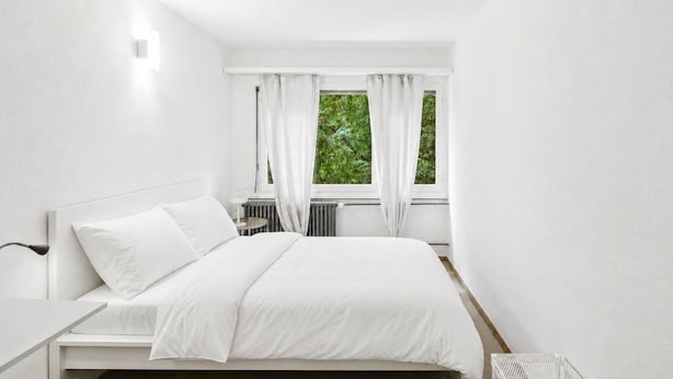 A bedroom with a bed, bedside table, and a window with curtains. - Sigma Self Check-In Hostel (Zürich)