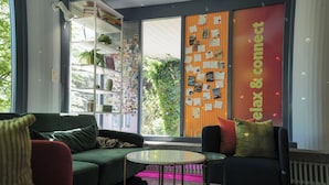 A modern room with a green sofa, a small round table, and a chair with colorful cushions. There is a large window with a view of greenery outside. - Sigma Self Check-In Hostel (Zürich)