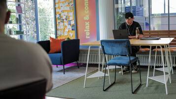 Eine Person arbeitet an einem Laptop an einem Tisch in einem Raum mit einer Pinnwand, Stühlen und einem Fenster.