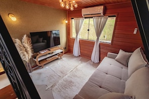 Ein gemütliches Wohnzimmer mit Flachbildfernseher, Holzmöbeln, einem großen Fenster mit Vorhängen und einer Deckenleuchte.