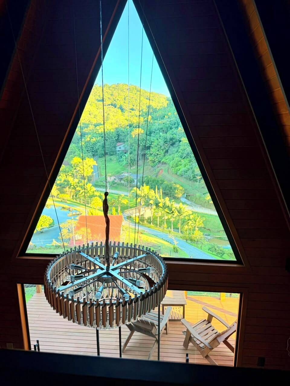 Ventana triangular con vistas a un paisaje verde, lámpara colgante circular y terraza de madera con sillas.