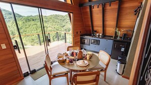 Un interior de madera con una mesa de comedor lista para cuatro comensales, una cocina con electrodomésticos modernos y una vista panorámica a través de amplias ventanas.