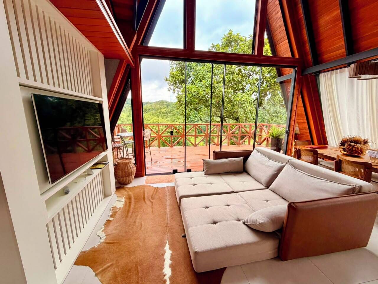 A modern living room with a large sectional sofa, a flat-screen TV mounted on a white wall, and a large glass door leading to an outdoor area with a wooden deck and greenery.