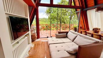 A modern living room with a large sectional sofa, a flat-screen TV mounted on a white wall, and a large glass door leading to an outdoor area with a wooden deck and greenery.