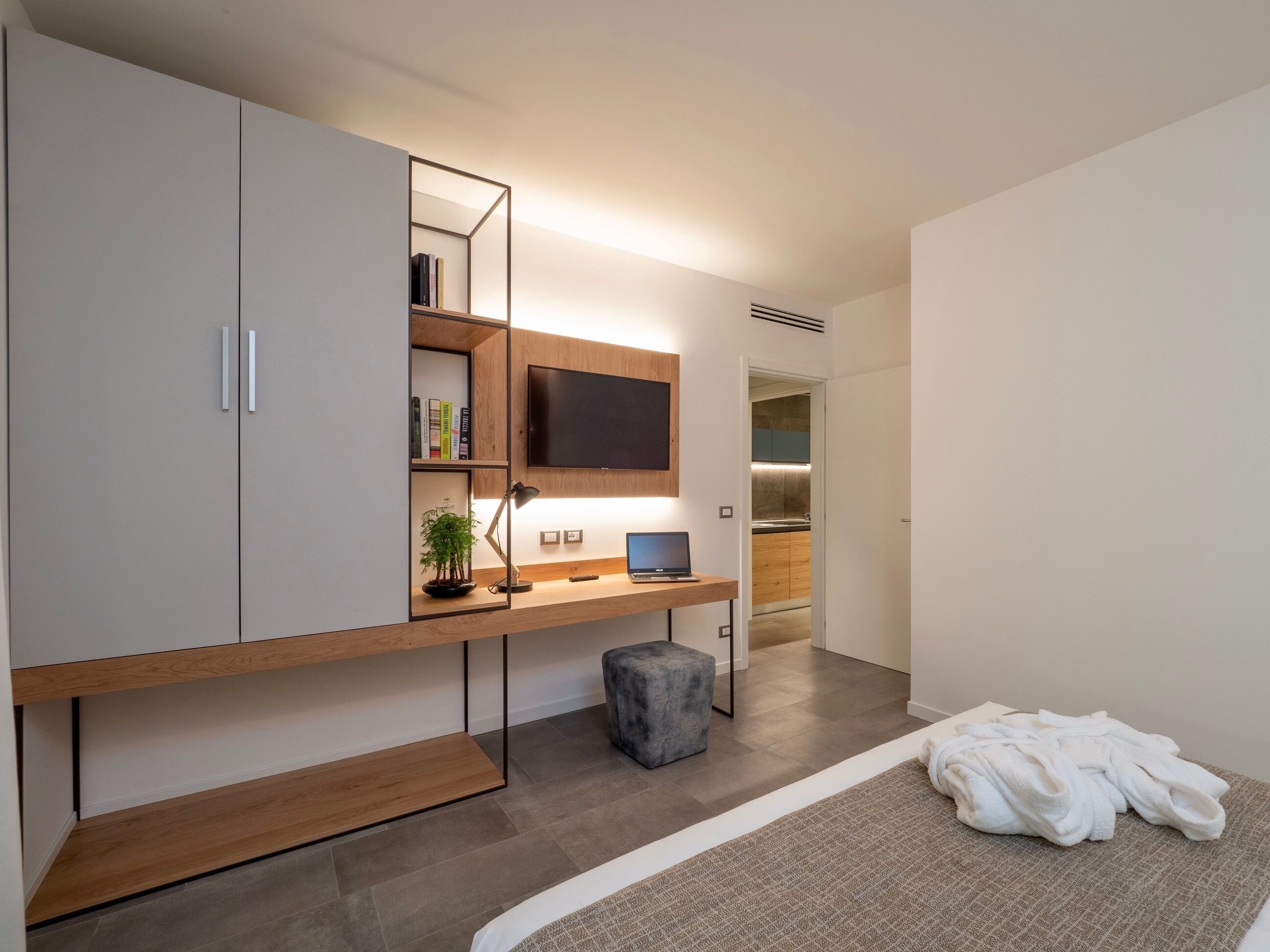 A modern bedroom with a large wardrobe, a desk with a laptop, a TV, and a bedside table with a lamp.