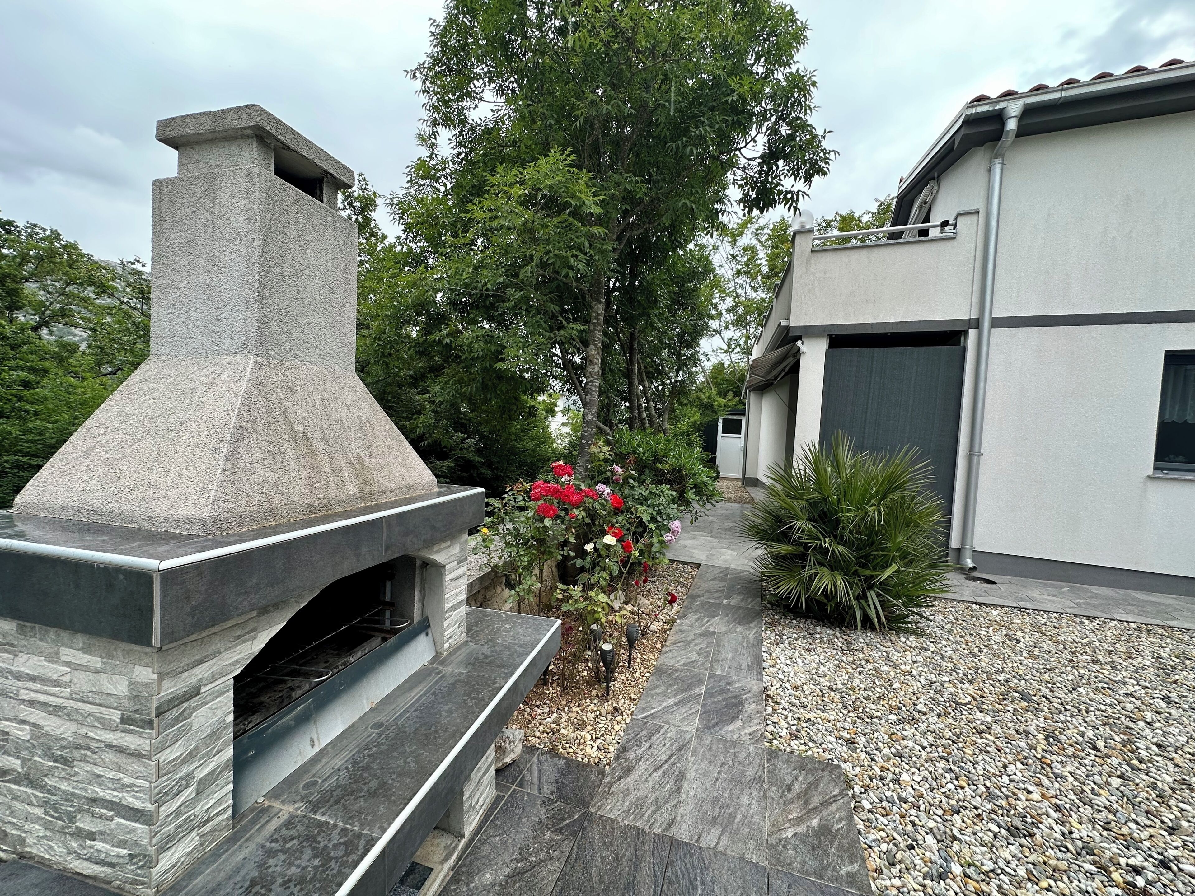 A modern outdoor kitchen with a stone chimney, surrounded by a garden with red flowers and a gravel pathway.