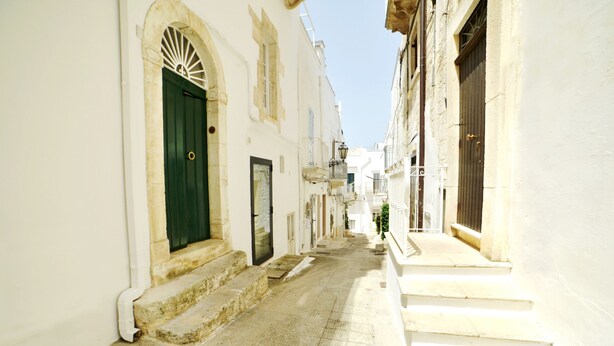 Una stretta via fiancheggiata da edifici bianchi, con una porta verde e scale che conducono a un balcone.