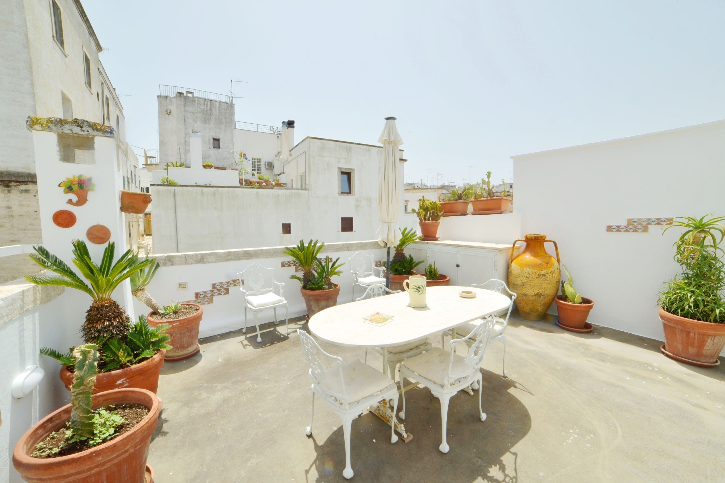 A rooftop terrace with a white table and chairs, surrounded by terracotta pots with various plants.