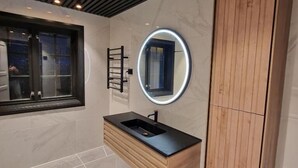 A modern bathroom with a wooden vanity, a round mirror with LED lighting, and a window with a view of the outside. - Aarnes House Stryn (Stryn)