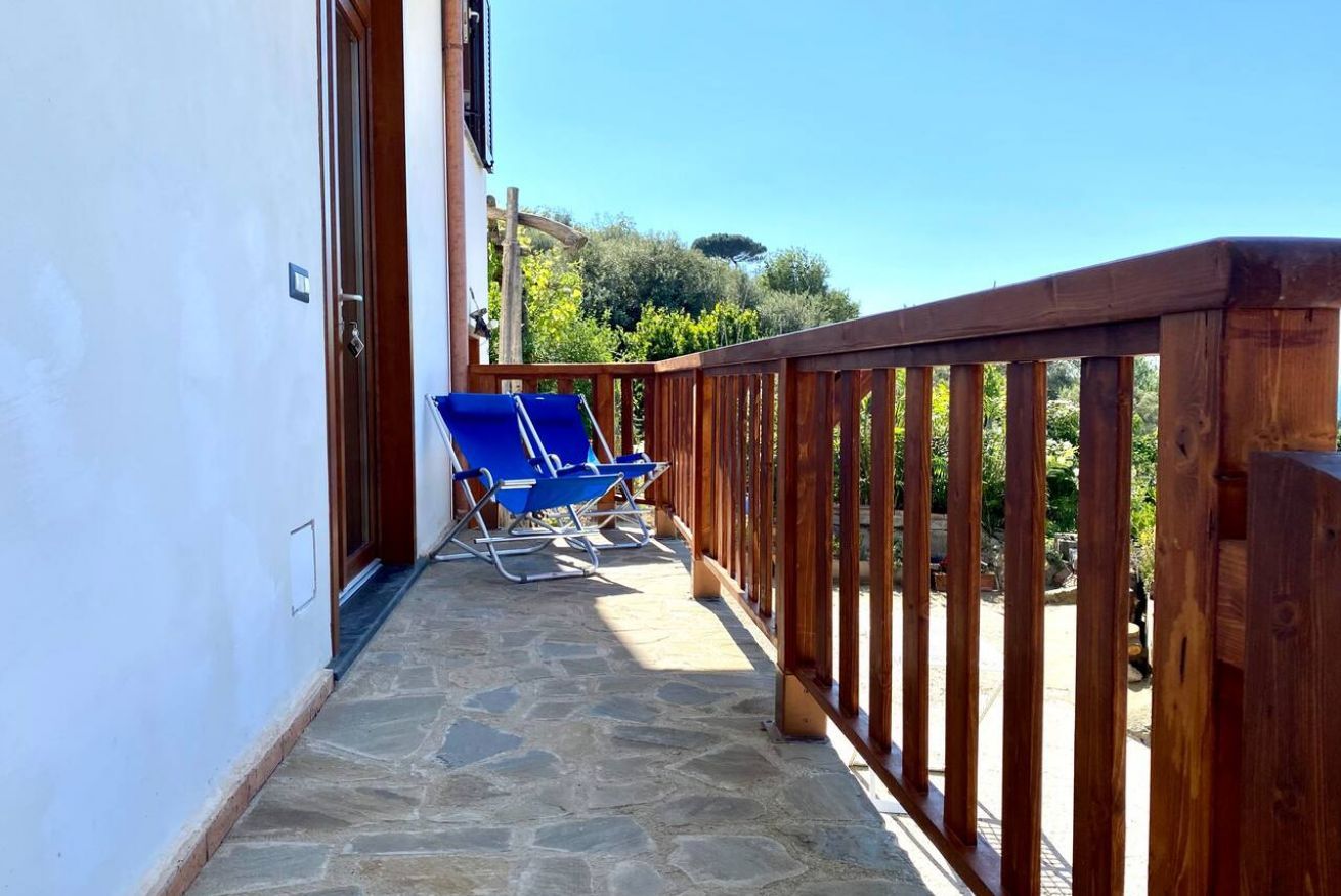 A stone-paved balcony with a wooden railing, two blue lounge chairs, and a clear view of greenery and trees in the background.