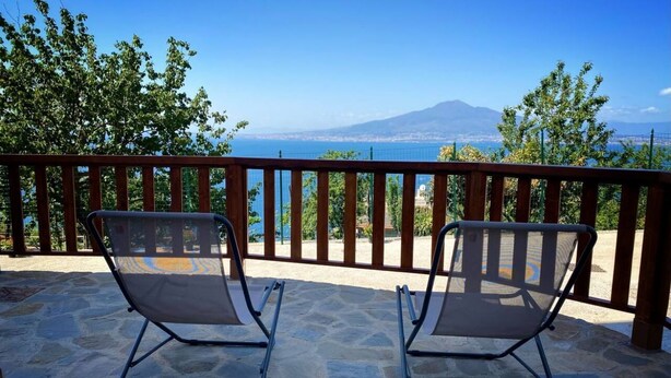 Two metal-framed lounge chairs on a stone patio with a view of a body of water and a distant mountain. - La Badia – Il Vigneto🌿 | Breathtaking View of Vesuvius and the Gulf of Naples (Vico Equense)