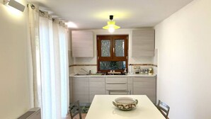 A kitchen with a white table, a window with curtains, and a ceiling light. - La Badia – Il Vigneto🌿 | Breathtaking View of Vesuvius and the Gulf of Naples (Vico Equense)