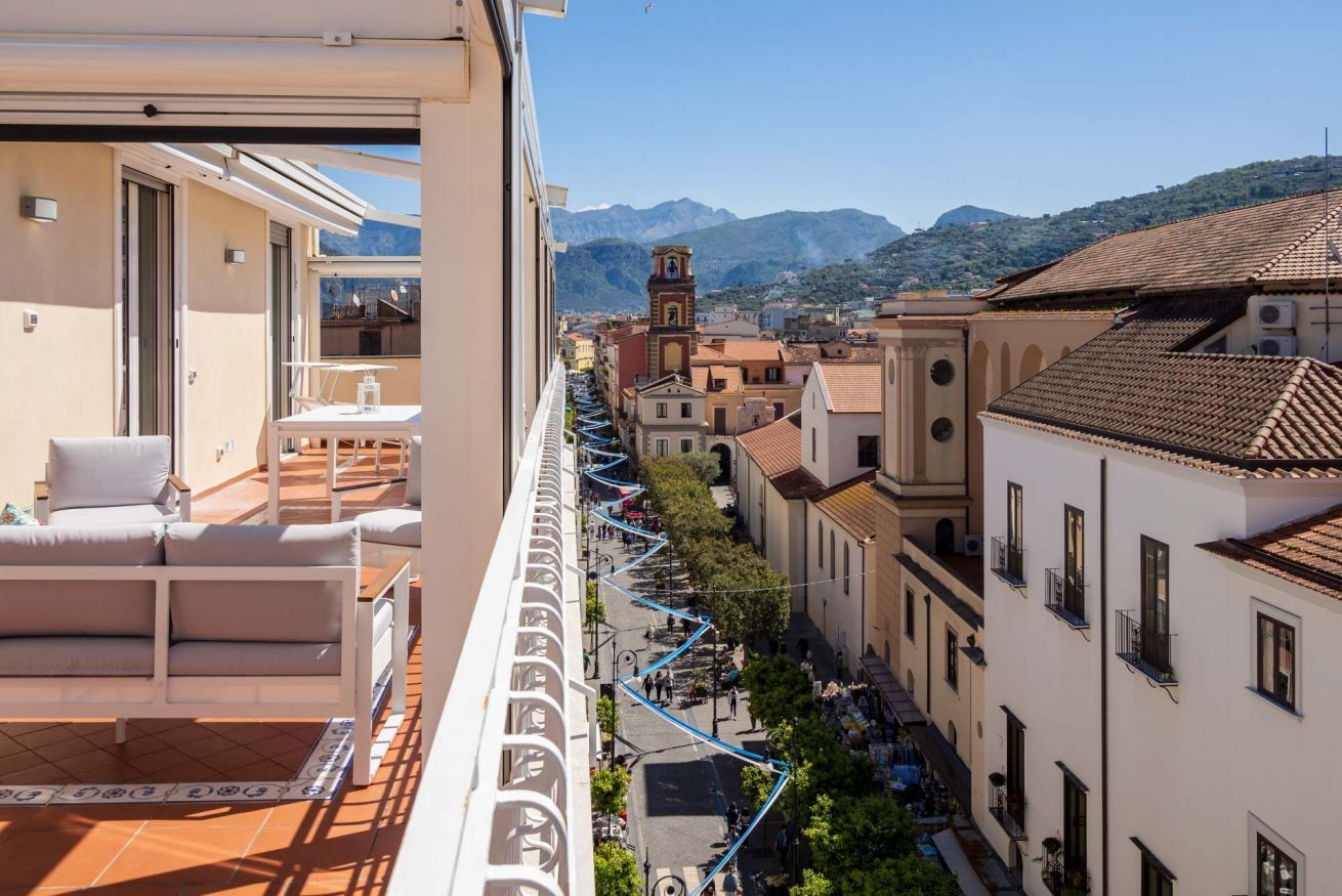 Un balcone con una divano e un tavolo, affaccia su una strada con edifici e una montagna sullo sfondo.
