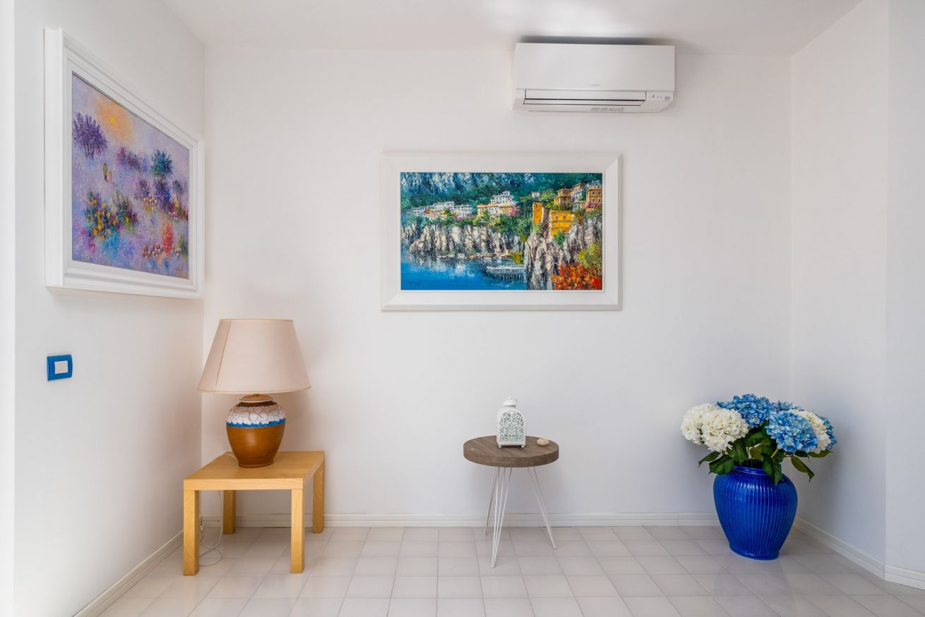 Quarto com parede branca, ar condicionado, um quadro, uma luminária, uma mesinha com uma vela e um vaso com flores.