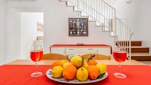Une table à manger avec une assiette de fruits et deux verres de vin rouge, un escalier et un tableau encadré au mur.