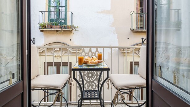 Un balcon avec deux chaises, une table où sont disposés des pâtisseries et une boisson, et une vue sur les bâtiments voisins.