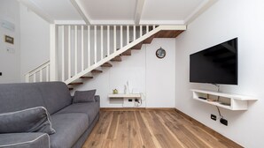 A modern living room with a grey sofa, wooden flooring, a white staircase, a mounted TV, and a small white shelf. - Casa Mirty – Comfort and the perfect location in the heart of the Sorrento Coast (Sant'Agnello)
