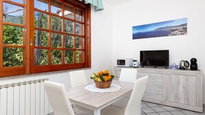 A dining area with a table, chairs, a fruit basket, a television, and a view of greenery through a window.