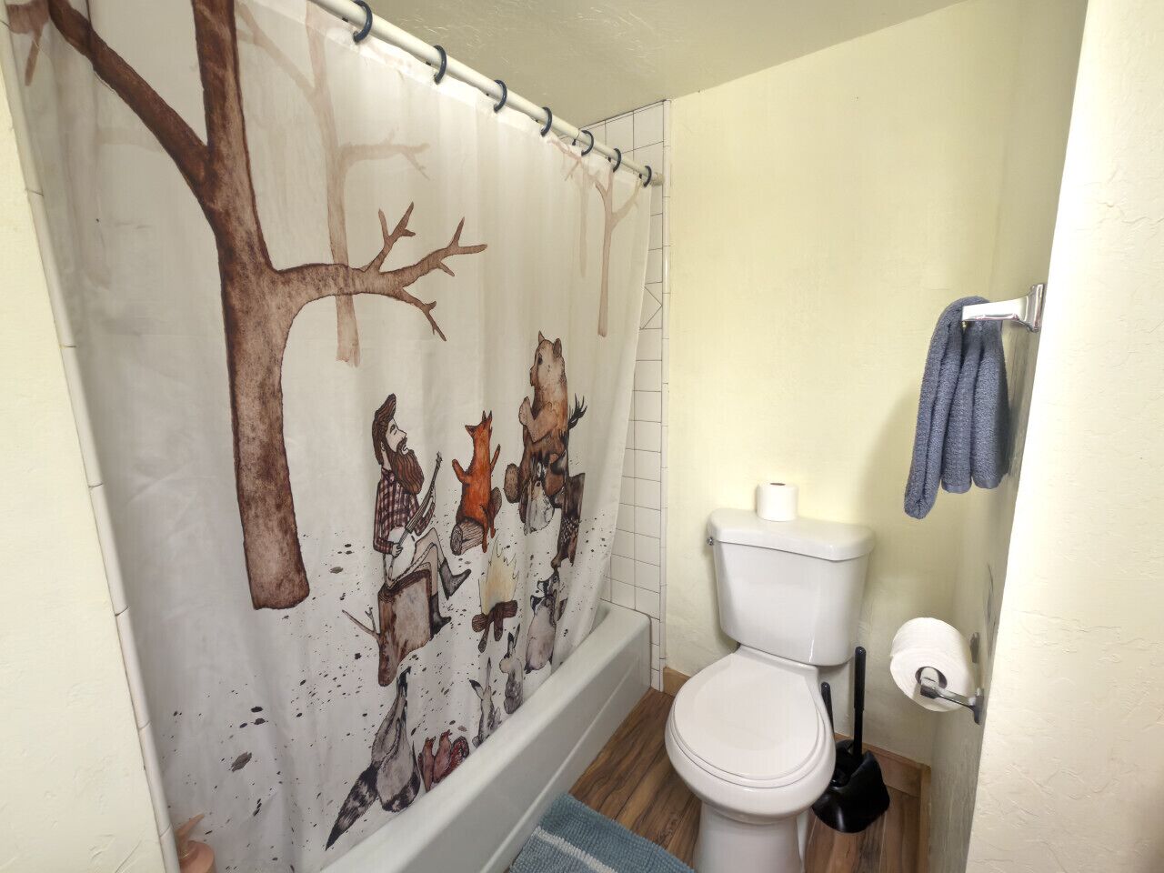 Une salle de bain avec un rideau de douche orné de motifs de cerfs et d’arbres, des toilettes et une serviette bleue accrochée au mur.