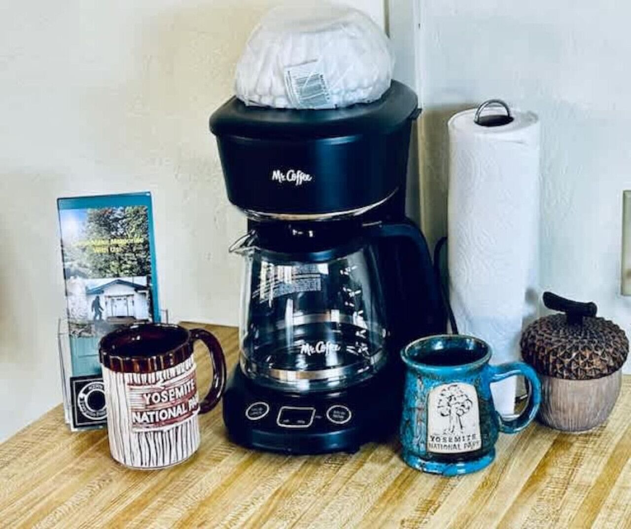 A coffee maker, two coffee mugs, a roll of paper towels, and a framed picture on a wooden surface.
