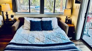 Une chambre à coucher avec un lit, deux tables de chevet, une fenêtre avec des rideaux et une vue sur des arbres et une voiture à l’extérieur.