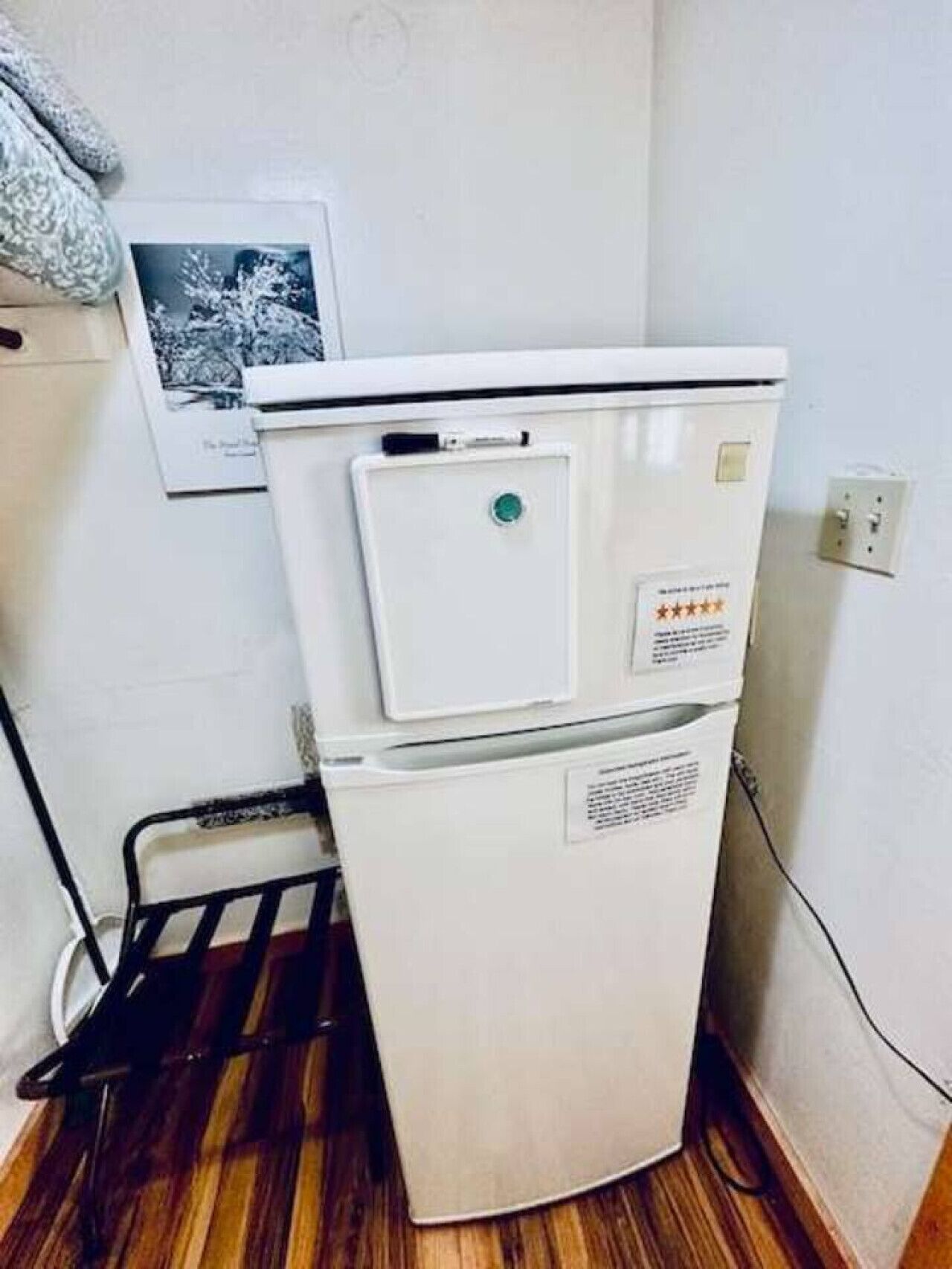 A compact refrigerator in a corner of a room with wooden flooring and a white wall.