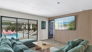 Ett modernt vardagsrum med en stor skjutdörr i glas som leder ut till poolområdet, ett trägolv och ett stort fönster med utsikt över stranden.