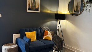 Una sala de estar moderna con un sofá azul oscuro, un reposapiés redondo de mimbre, una alfombra estampada y una lámpara de pie.