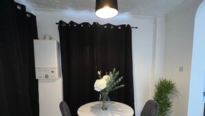 Un comedor con una mesa redonda, dos sillas, un florero con flores, una cortina negra, una pared blanca, una lámpara de techo y una planta en maceta.