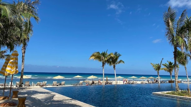 A poolside area with lounge chairs and umbrellas, palm trees, and a view of the ocean.