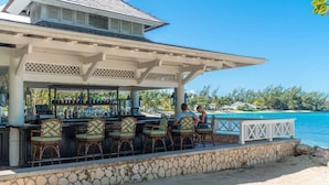 A beachside bar with stone walls and wicker chairs, overlooking a clear blue ocean and palm trees.