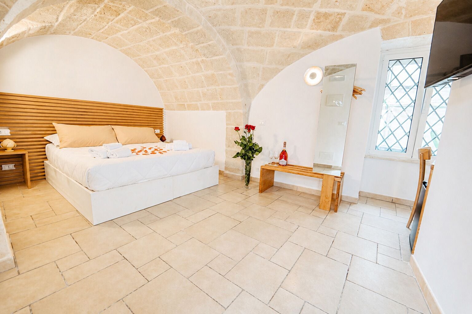 Une chambre à coucher avec un lit, un banc en bois, un vase de fleurs, un miroir et un téléviseur.