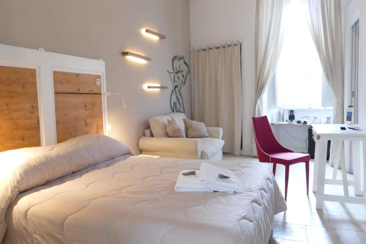 A bedroom with a large bed, a desk, and a chair. There are wall-mounted lights and a window with curtains.
