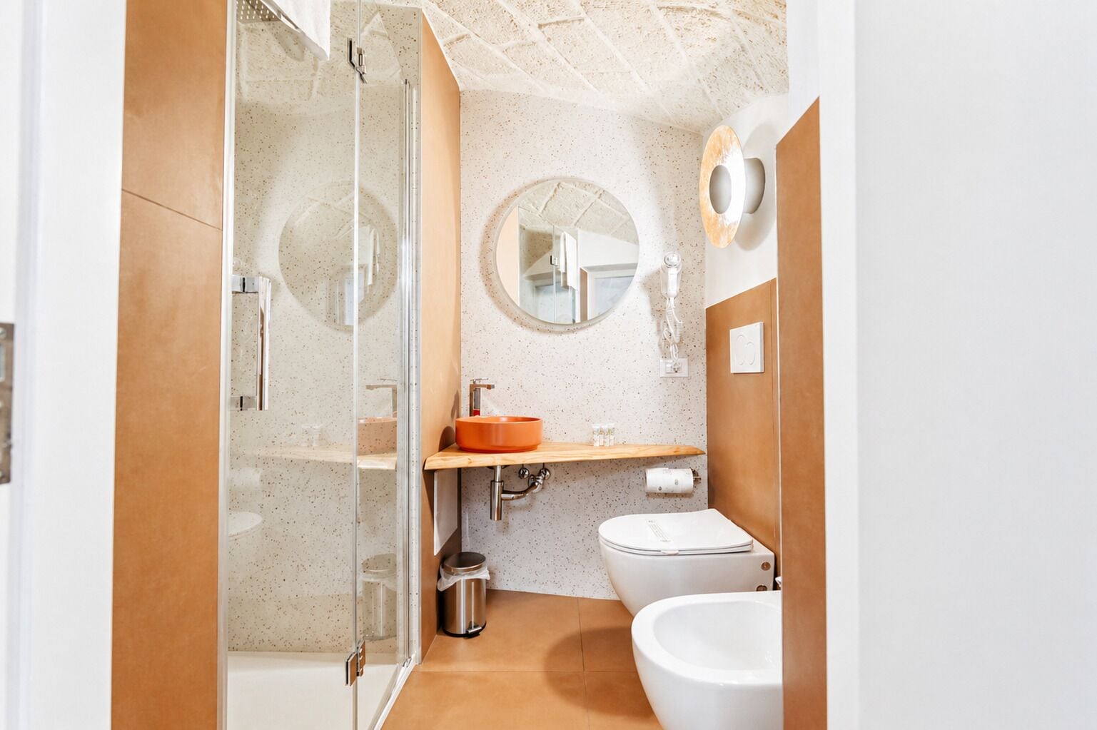 Un baño moderno con cabina de ducha de vidrio, un espejo redondo, tocador de madera y un inodoro blanco.