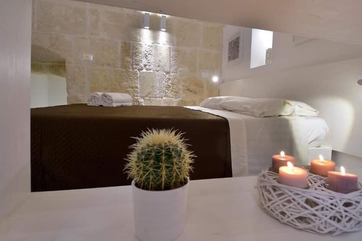 A bedroom with a stone wall, a bed with white linens, a cactus in a white pot, and three lit candles in a woven basket.