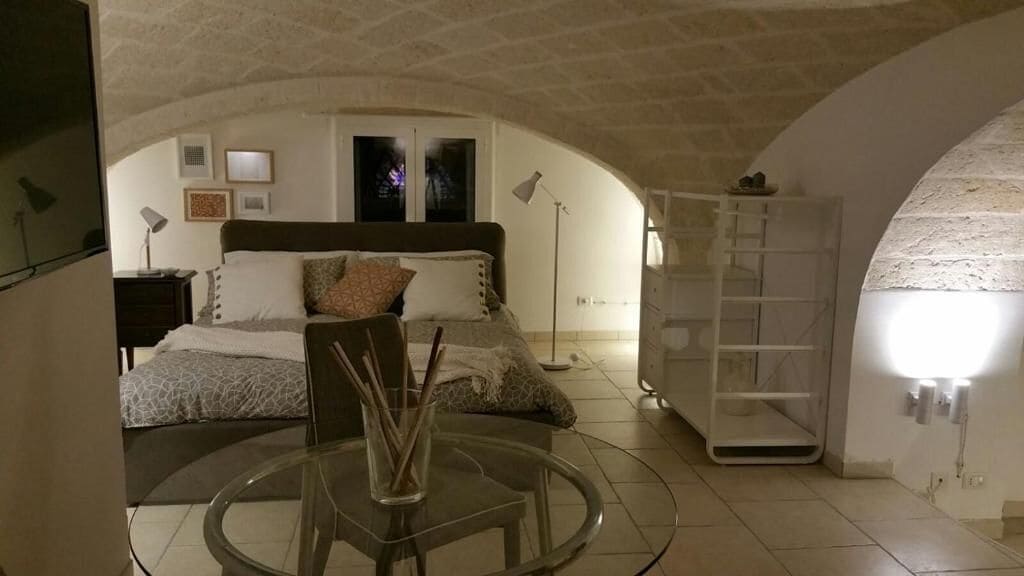 A bedroom with a bed, bedside table, lamp, and a glass table with a vase.