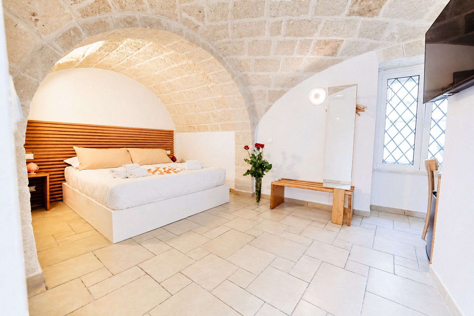 Ein Schlafzimmer mit Steinbogen-Decke, einem Bett, einer Bank, einer Vase mit Blumen und einem Fernseher.