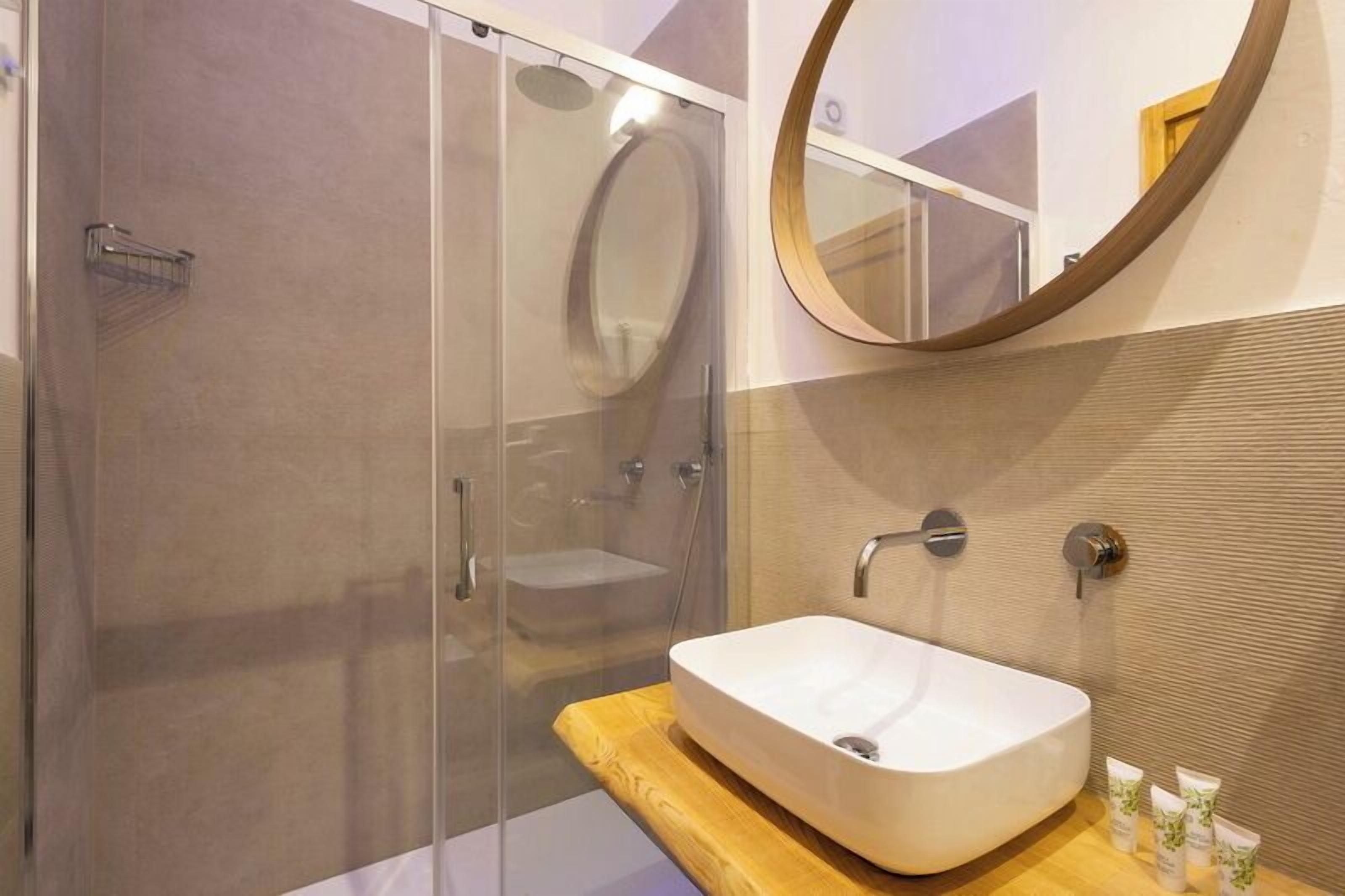 Een moderne badkamer met een douche met glazen wand, een vrijstaand badkamermeubel en een ronde spiegel.