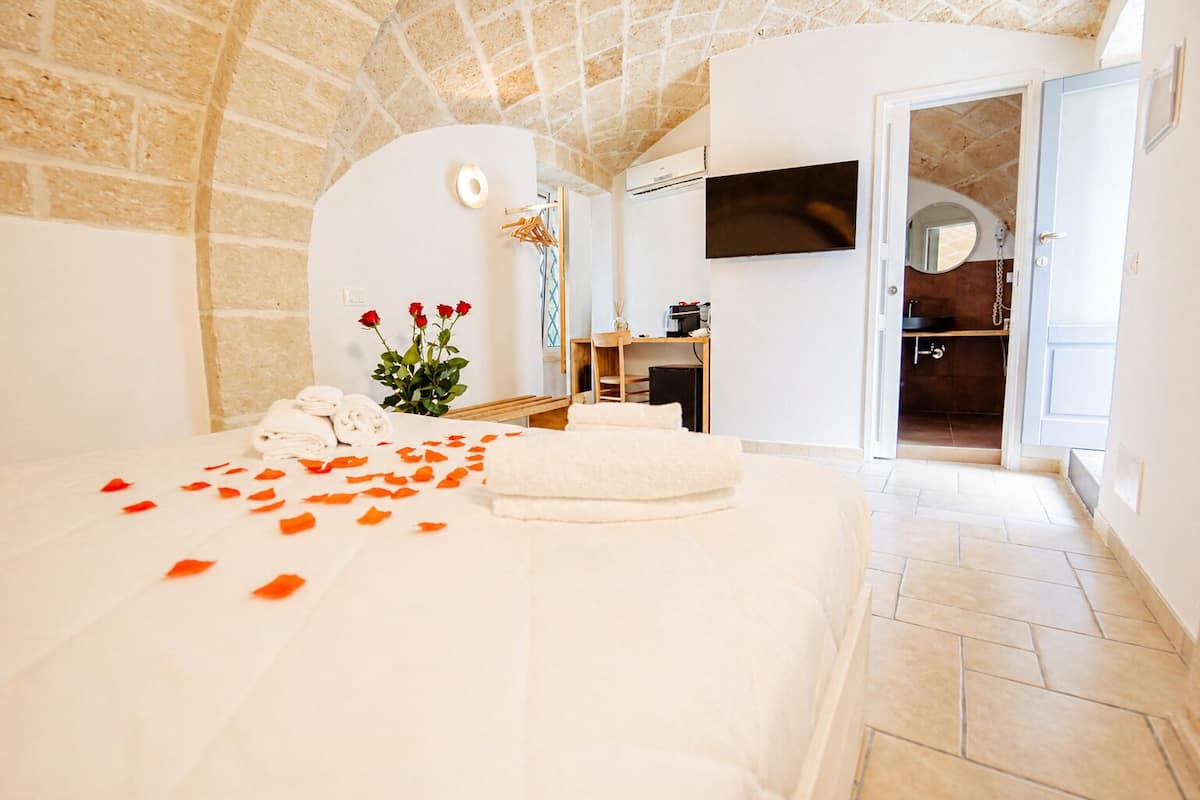 A hotel room with a stone ceiling, a bed with white linens, a TV on the wall, and a bathroom visible through an open door.