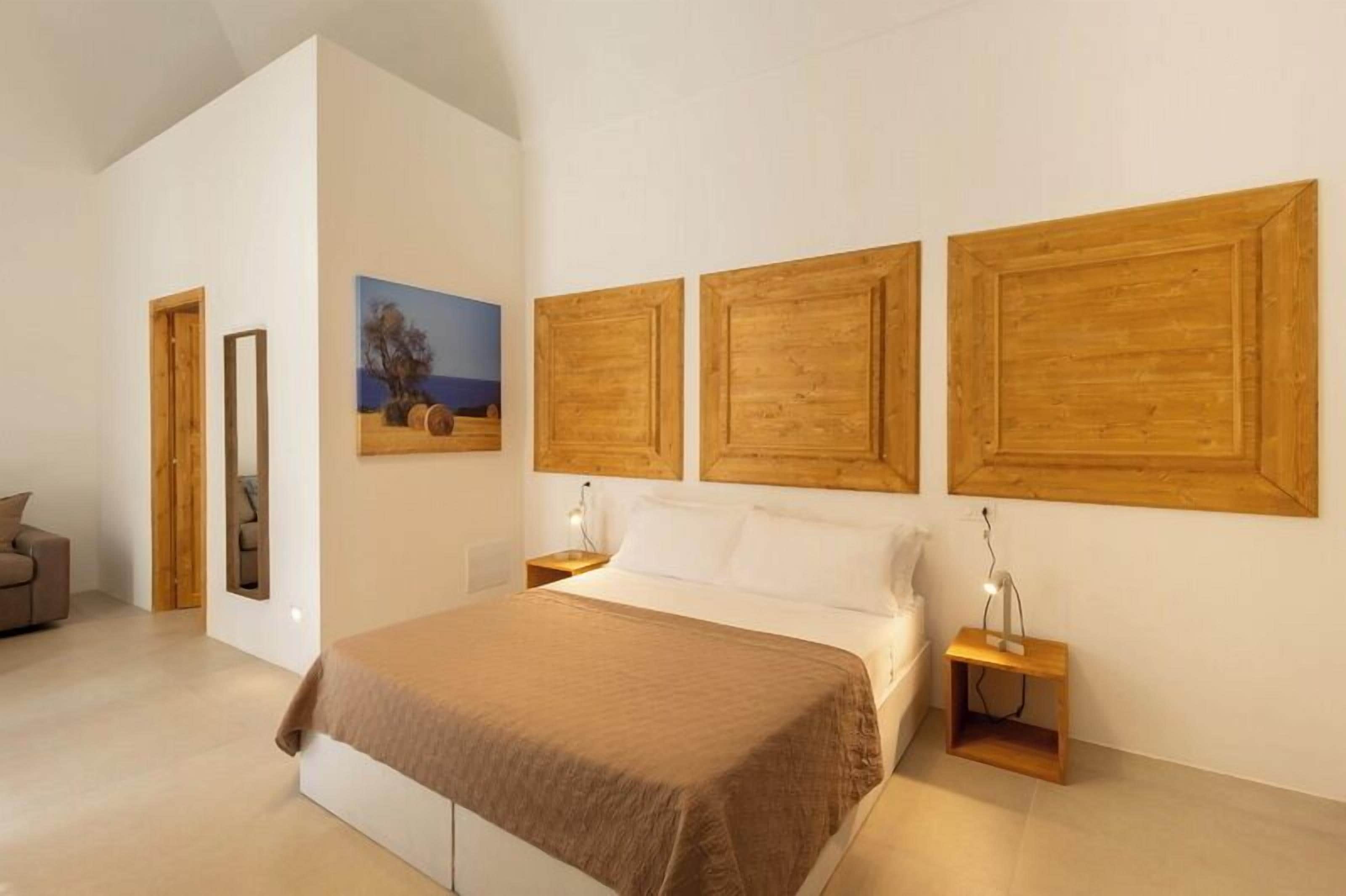 A modern bedroom with a large bed, wooden headboards, a painting, and a small bedside table with a lamp.